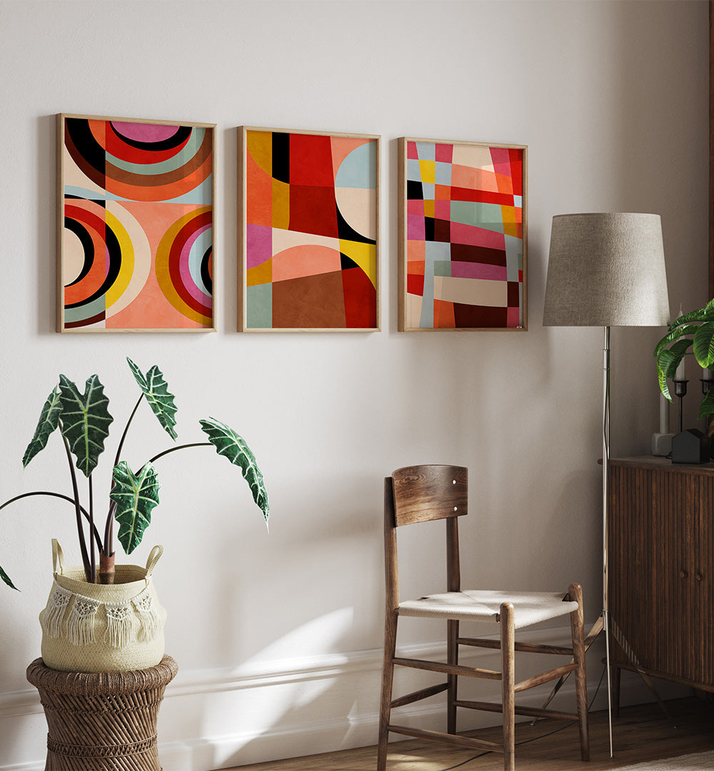 Three abstract art pieces on a wall with a chair, plant, and lamp in a room.