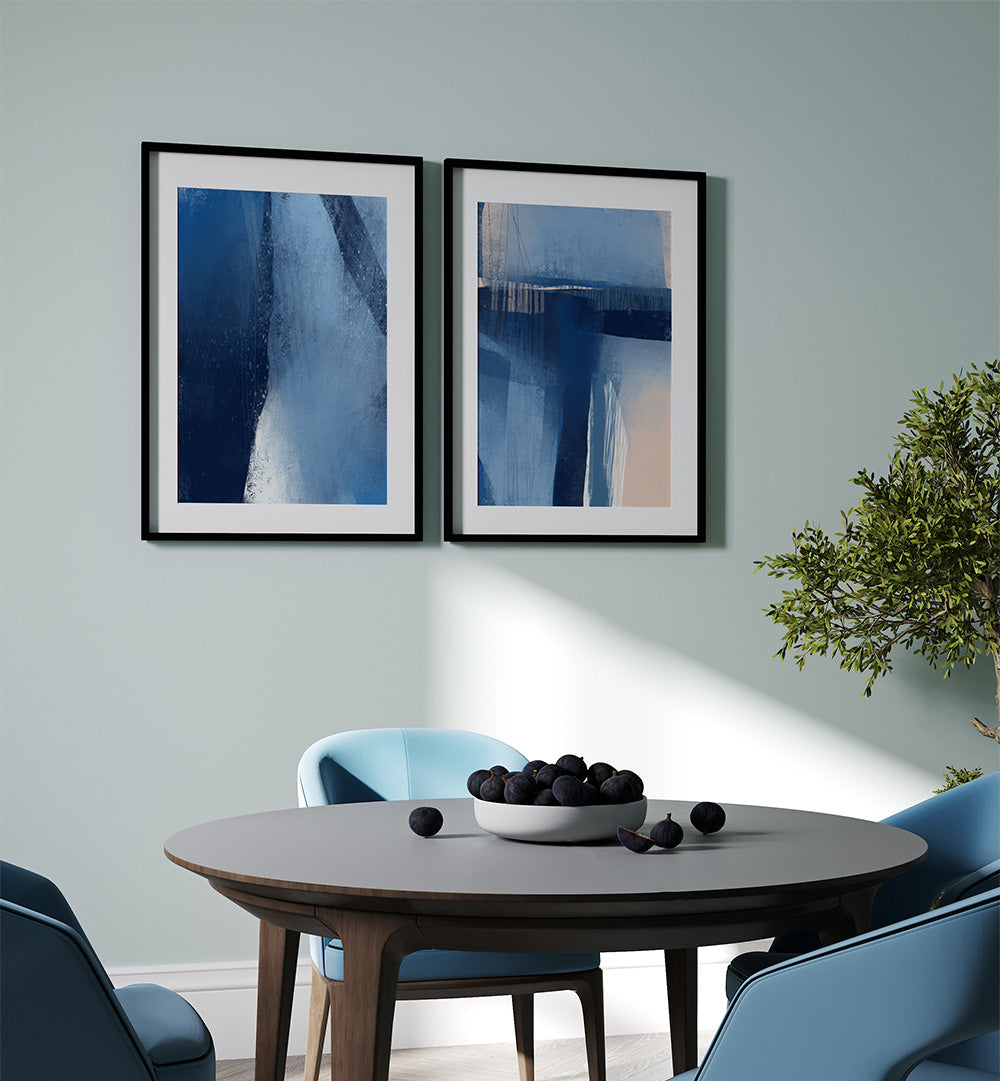 Two abstract paintings on a wall above a round wooden table with a bowl of fruit.