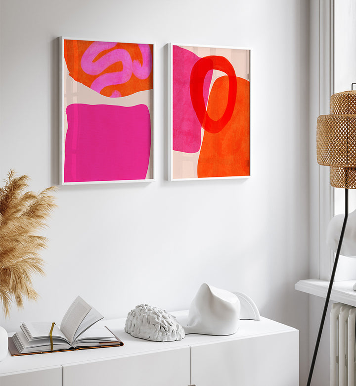 Two abstract paintings with pink and orange colors on a white wall above a white cabinet.