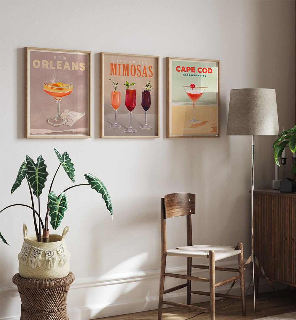 Three framed cocktail posters on a wall with a chair, lamp, and plant in the foreground.