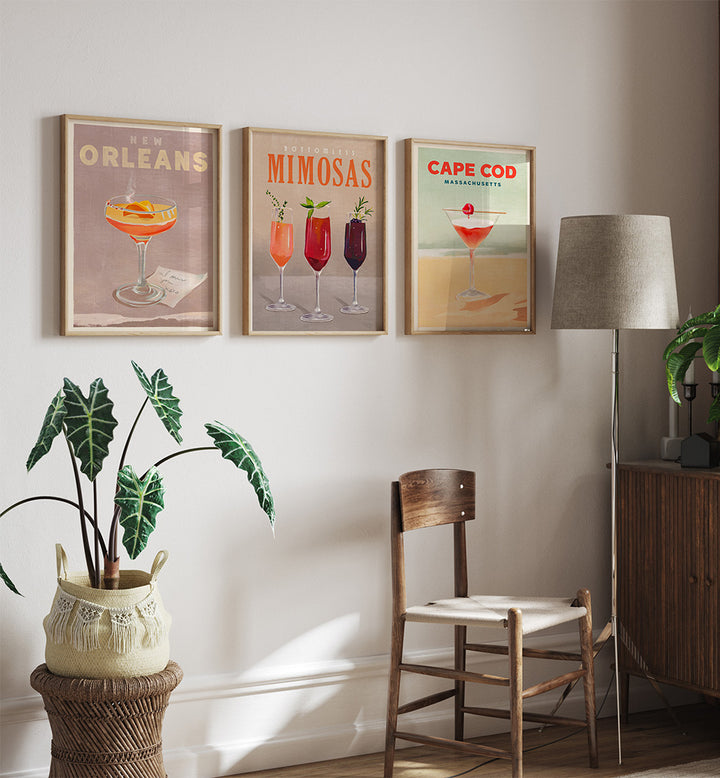 Three framed cocktail posters on a wall with a chair, lamp, and plant in the foreground.