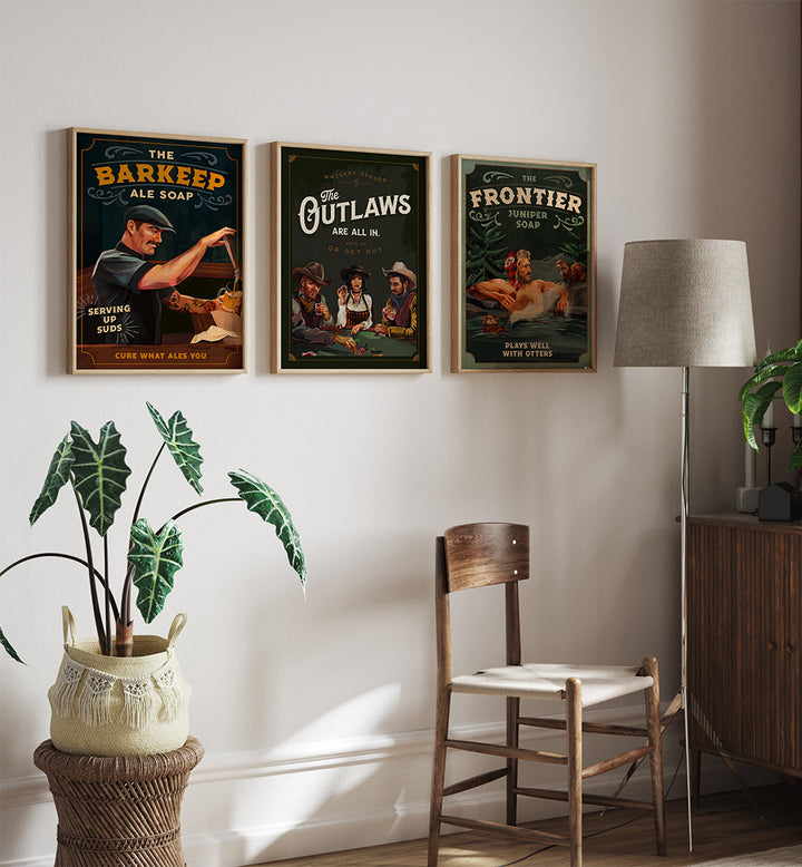 Three framed posters on a wall above a wooden chair with a lamp and plant in the room.