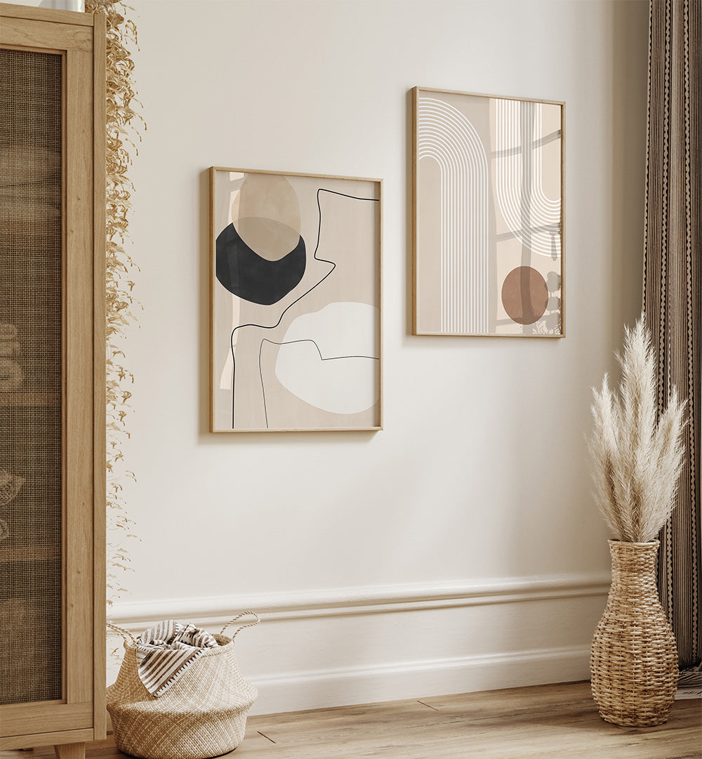 Two abstract art pieces on a wall with a vase of dried plants and a basket below.