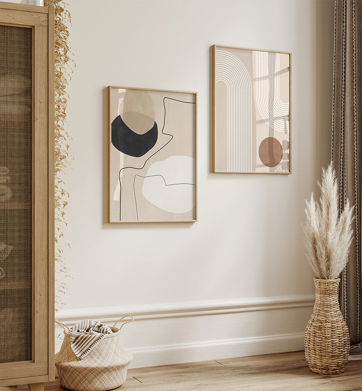 Two abstract art pieces on a wall with a vase of dried plants and a basket below.