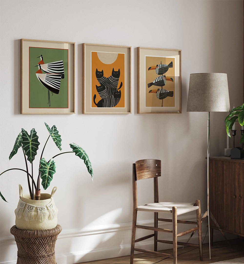 Three framed artworks on a wall above a wooden chair with a plant and lamp in the room.