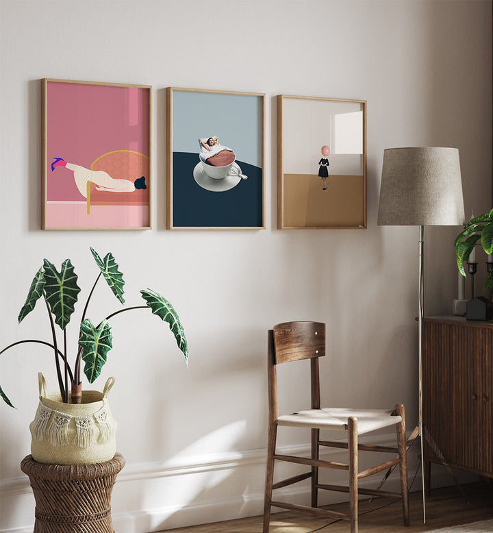 Three framed artworks on a wall with a chair, lamp, and plant in a room.