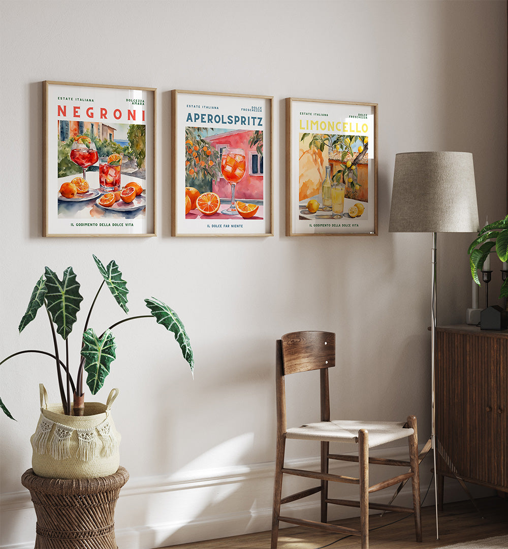 Three framed posters on a wall above a wooden chair with a plant and lamp in the room.