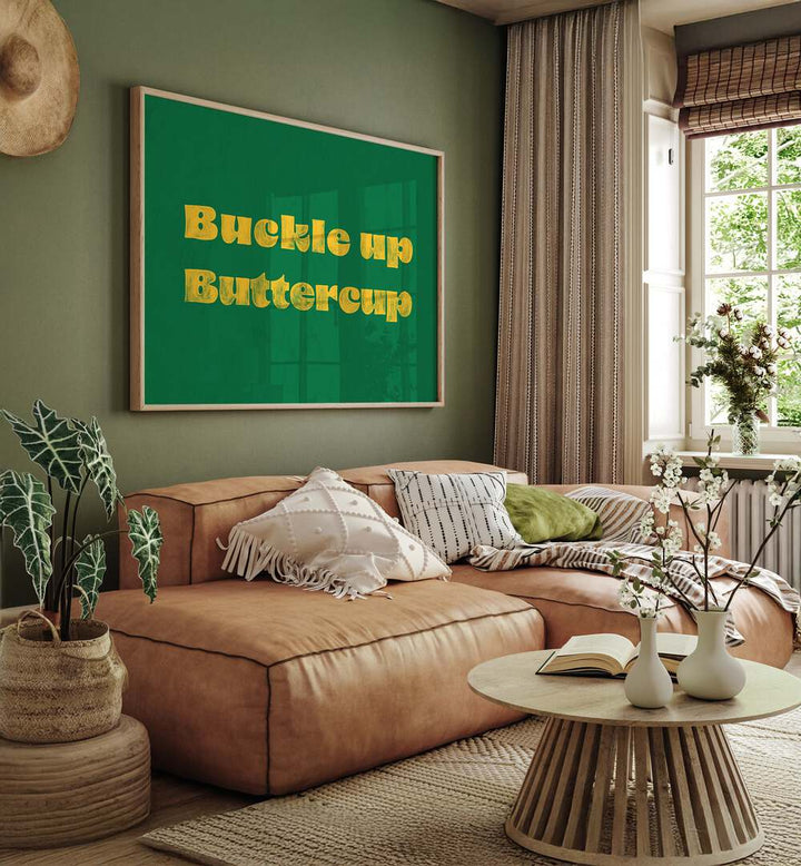 Buckle Up Buttercup Quotes And Typography Posters in Oak Wood Plain Frame placed on a Green Colored Wall near a Brown Sofa in the Living Room