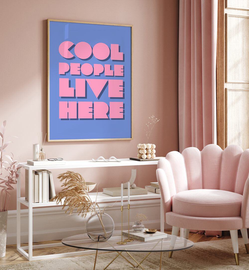 Cool People Live Here by Athene Fritsch Quotes and Typography Posters in Oak Wood Plain Frame placed on a pink wall beside a window and behind a table