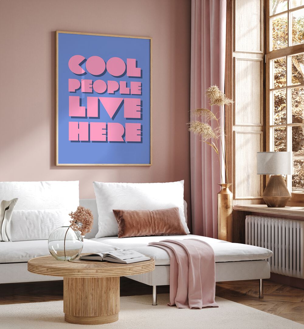 Cool People Live Here by Athene Fritsch Quotes and Typography Posters in Oak Wood Plain Frame placed on a pink wall beside a window and behind a sofa for living room