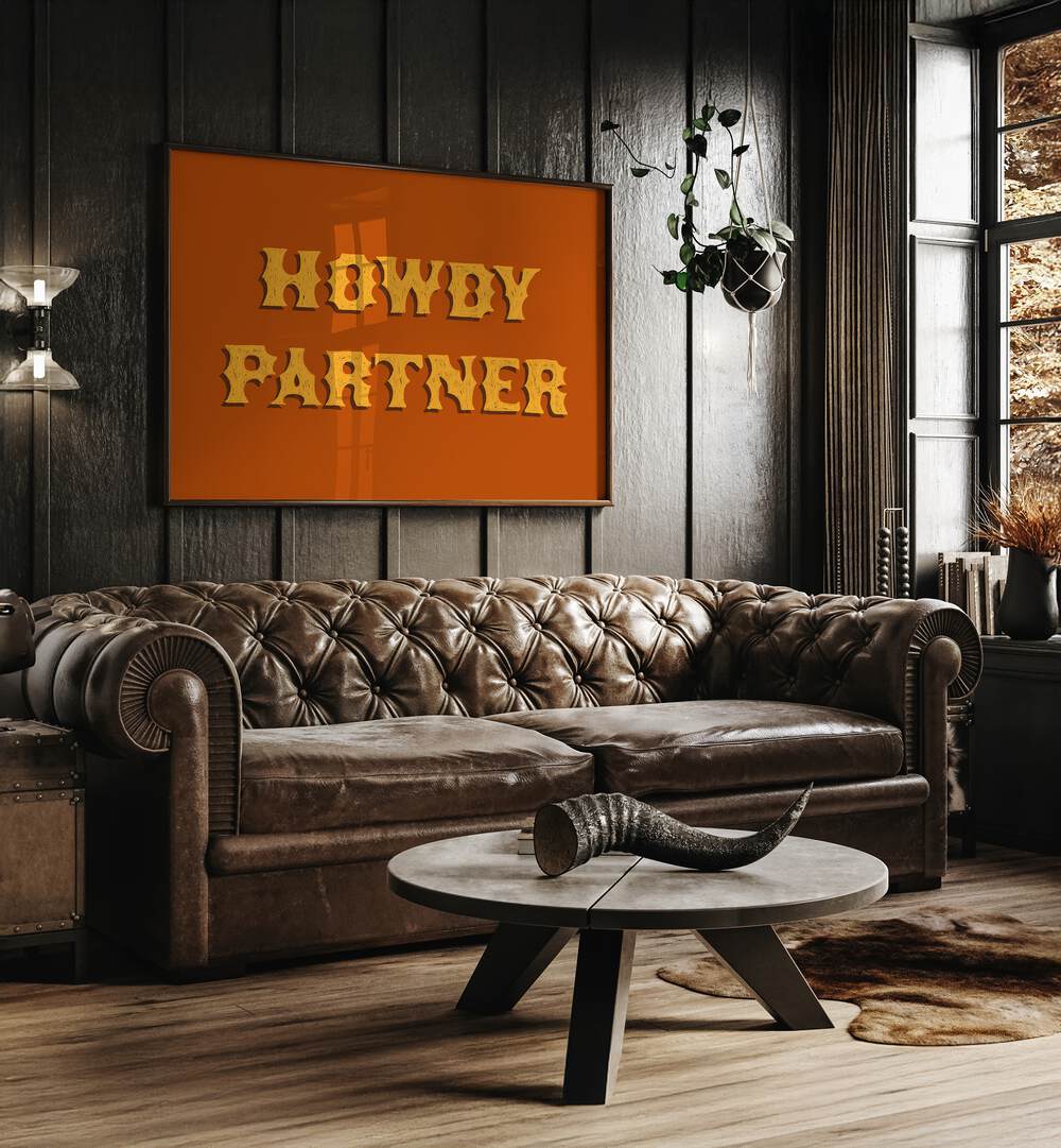 Howdy Partner Quotes And Typography Posters in Dark Wood Plain Frame placed on a Wooden Textured Wall near a Brown Leather Sofa in the Living Room