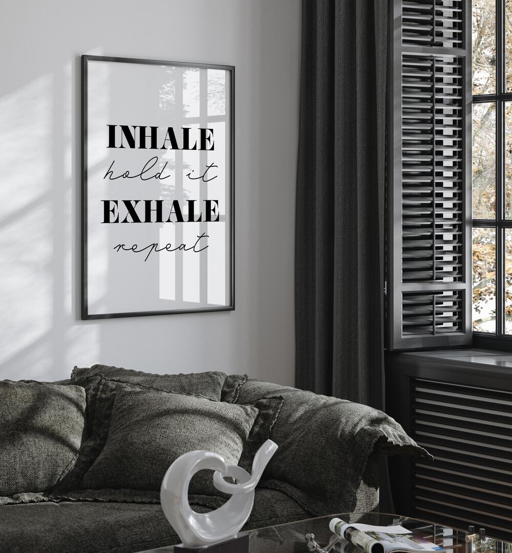 Inhale And Exhale By 1 X Studio Quotes And Typography Posters in Black Plain Frame placed on a White Colored Wall near a Dark Grey Sofa in the Living Room