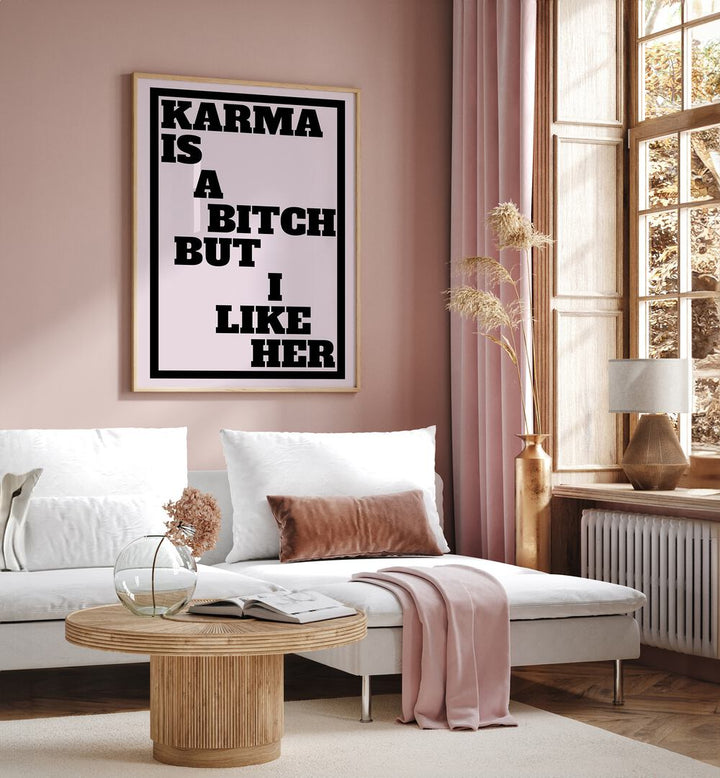 Karma is a Bitch by Athene Fritsch Quotes and Typography Posters in Oak Wood Plain Frame placed on a pink wall beside a window and behind a sofa for living room