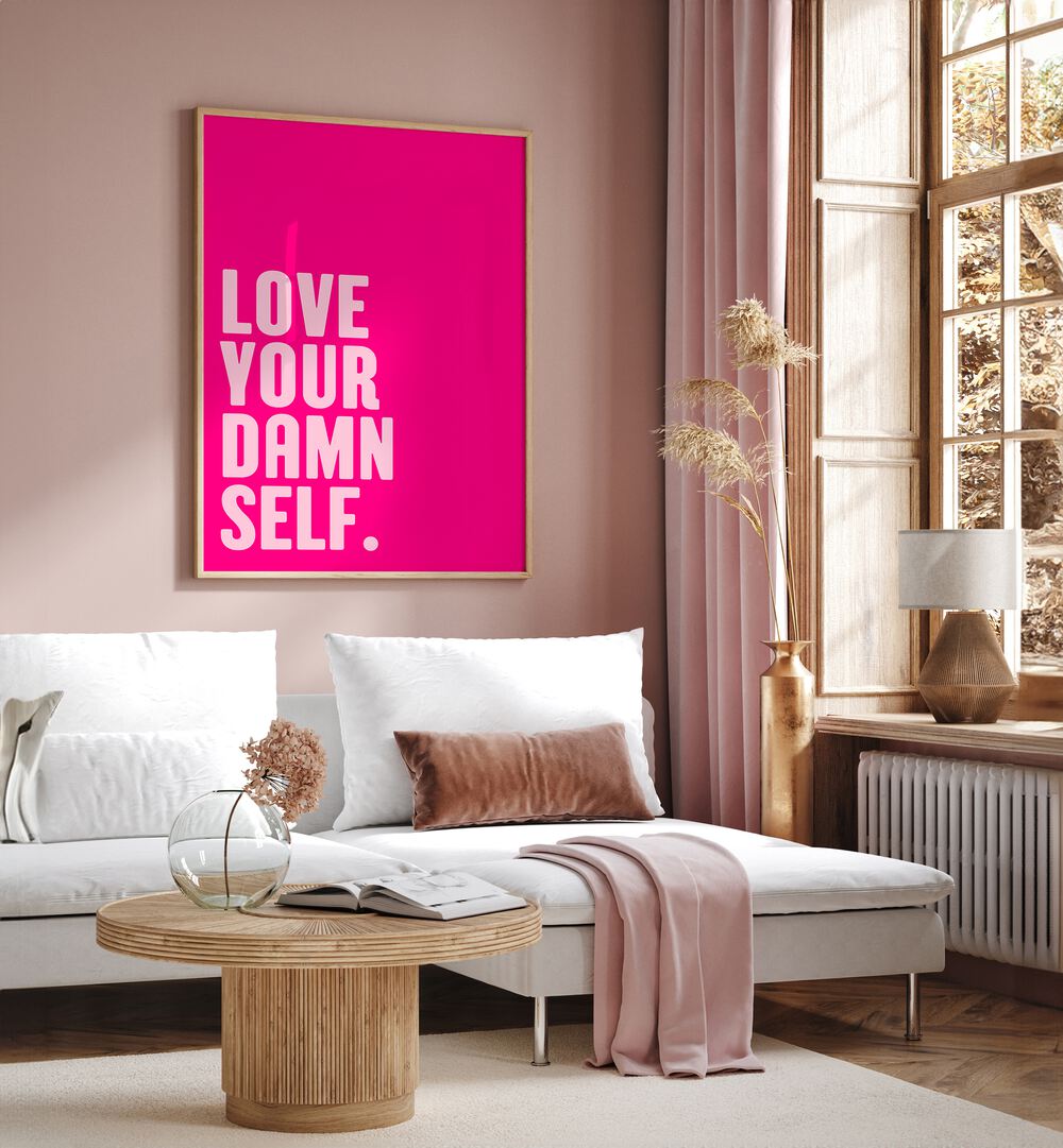 Love Your Damn Self by Athene Fritsch Quotes and Typography Posters in Oak Wood Plain Frame placed on a pink wall beside a window and behind a sofa for living room