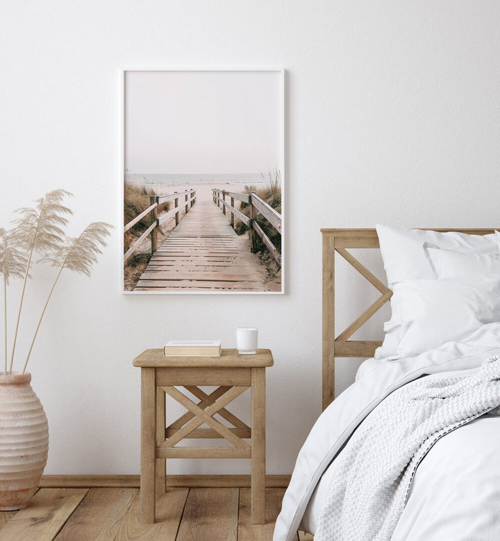 Sandy Promenade Beach Prints Coastal Wall Art in White Plain Frame placed on a White Colored Wall near a Bed in the Bedroom