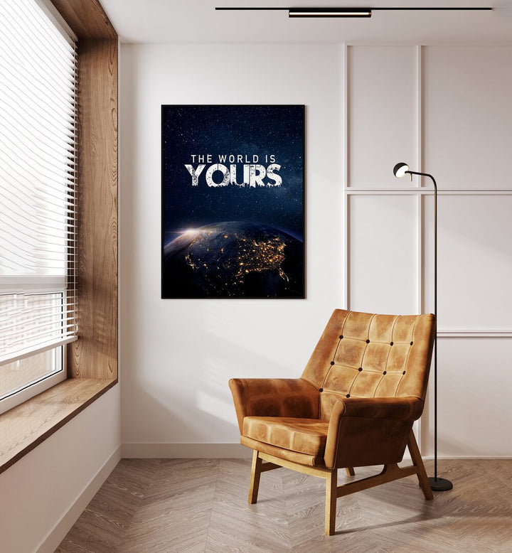 The World Is Yours Quotes And Typography Posters in Black Plain Frame placed on a Cream Colored Wall near a Brown Sofa Chair in the Drawing Room