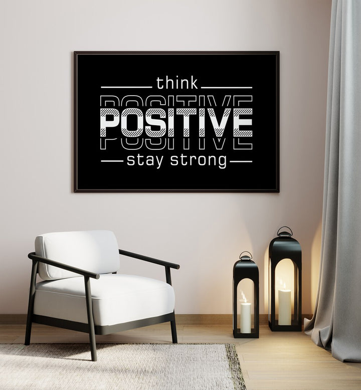 Think Positive Stay Strong Quotes and Typography Posters in Black Plain Frame placed on a wall in a room beside a chair